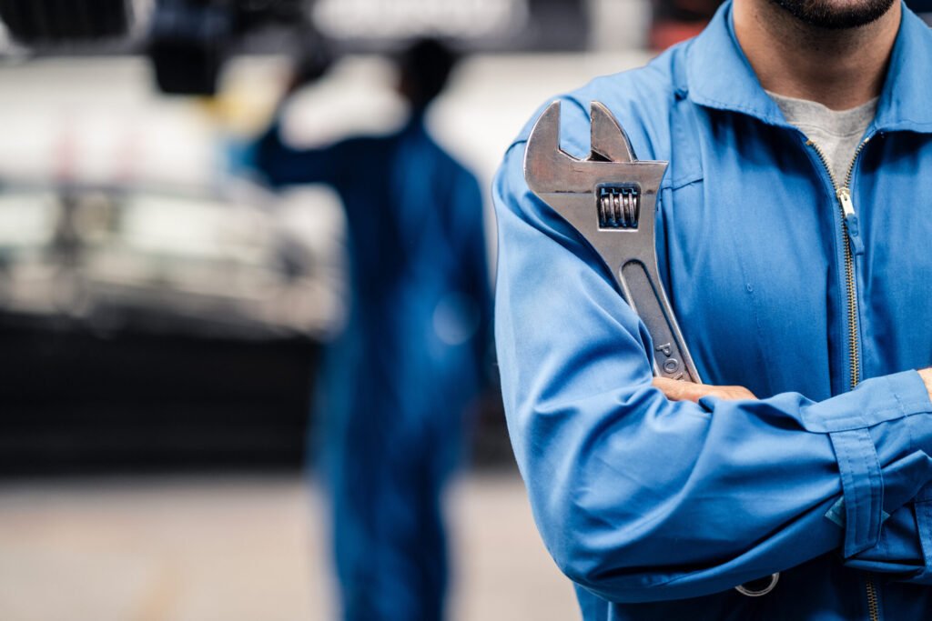 Man in overalls crossed arms with an adjustable spanner, colleage working in the background